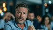 © pattozher - Anxious Business Meeting: A middle-aged man looks distressed and worried in a dimly lit office meeting, with blurred figures in the background expressing concern.