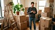 © Monera - Smiling man in casual sweater tapping plain smartphone surrounded by stacked cardboard moving boxes in sunlit apartment with wooden ladder, houseplants, and bicycle
