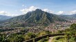 © Tandu - Panoramic view of a sprawling city nestled beside majestic green mountains under a bright blue sky with scattered clouds.