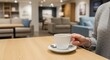 © Lee - Hand holding a warm white coffee cup and saucer on a wooden table with a blurred interior background