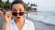 © benjamin - A woman with a shocked expression on her face peers over sunglasses on a tropical beach, with gentle waves lapping at the shore and palm trees in the background.