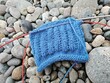 © Elena - Blue knitted swatch on red circular needles resting on smooth river stones. Handmade craft and knitting hobby concept