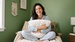 © haris - A smiling woman in striped pajamas sits cross-legged on a wooden chair, holding a white pillow in a cozy room with green walls.