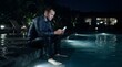 © Danawan Purbanggoro - A soaked businessman in a dark shirt sitting on the edge of an illuminated swimming pool at night while checking his smartphone with a serious look.