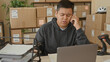 © Krakenimages.com - Man holds smartphone to ear and writes by laptop amid stacked parcel boxes and barcode scanner in a building; small business stress.