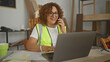 © Krakenimages.com - Woman carpenter holds phone to ear and types on laptop amid wood boards and safety glasses in a studio; focused planning.