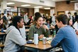 © Kassiopeia  - Three teenagers sit at a table in a school cafeteria. They are sharing food and laughing. Other students are in the background eating and chatting