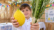 © olgakris - Large citron held toward camera by boy, child offering etrog beside bound lulav in a festive sukkah, festival promotion banner for seasonal faith gathering