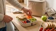 © Nusrat - Unidentified man packs nutritious lunch with whole grain sandwich carrot sticks grapes and mixed berries into bento box for healthy office meal prep.