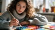 © Pete - Young woman paints with watercolor on table. Cozy knit sweater shows creative focus. Artist uses brush and palette inside bright home studio.