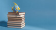 © RIMA - Blue and yellow fish figurine resting on a tall stack of books against a light blue background