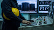 © NK - Industrial process control and plant monitoring concept, operator using a console keyboard while holding a yellow hard hat beside illuminated switches and gauge displays in a factory control room