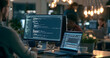 © Jack - A Closer Look at a Computer Monitor Displaying Backend Code and a SQL Database Query, alongside a Laptop, as an Indistinct Software Developer Writes Scripts in a Bustling Work Environment with Various