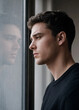 © SerPak - Young man with a thoughtful expression gazes through a window, his reflection visible in the glass, creating a quiet introspective portrait indoors.