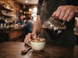 © Pravinrus - Close-up of barista pouring hot freshly brewed black coffee from a glass server into a ceramic cup on a wooden counter in a warm rustic cafe setting.