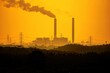© ANTOSYS - Telephoto view of industrial plant chimneys blowing thick smoke against a bright yellow sunset sky.