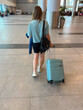 © Krystsina - Young female traveler in casual attire pulls a light blue suitcase through a modern airport terminal with spacious interior and travelers in the background