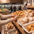 © Hafiz - Person selecting fresh artisan bread from a wooden shelf in a busy grocery store bakery with person reaches rustic loaf bread shelf filled various baked goods supermarket