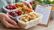 © ISVO - Individual holding three containers of frozen food including fruits, vegetables, and grains with a meal plan note visible in a bright kitchen setting