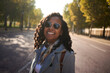 © CarlosBarquero - Cheerful African millennial in a gray coat and sunglasses smiles at the camera on a sunlit, tree-lined city street, with soft bokeh and autumn vibes suggesting travel and relaxed