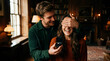 © DIGADO - Young man surprising his excited girlfriend by covering her eyes with his hands and holding an open box with a diamond ring for a romantic marriage proposal in a cozy, warm-lit living room interior