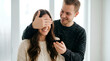 © DIGADO - Young happy man surprising his smiling girlfriend by covering her eyes with one hand while holding an open black engagement ring box during a romantic marriage proposal indoors near a bright window