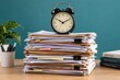 © Bakar Gallery - A black alarm clock sits atop a towering stack of cluttered paperwork on a wooden office desk. This image represents workplace time management and deadline stress concepts.