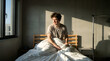 © Yuliia - Young man waking up in sunlit bedroom, stretching on bed with white blanket