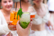 © dsbedarieva - Friends toast with cocktails including a mojito, spritz, and coupe drink in Corfu, Greece. A close-up shot highlights hands holding iced glasses at an outdoor table.