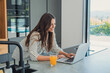 © Daniel - Woman smiling and typing on laptop computer, enjoying remote work from home in a contemporary kitchen with a glass of fresh orange juice on the counter