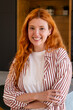 © simona - Confident redhead woman smiling in a casual striped shirt, portrait of a friendly young adult with crossed arms in a modern interior conveying warm, relaxed professionalism