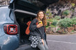© Koval988 - Young woman sits on the open trunk of a car, holding a phone. On a road trip with her phone and backpack, she enjoys the natural scenery outdoors. An active lifestyle.