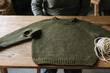 © Milos - An unfinished knitted sweater with a hole lies flat on a wooden table, showcasing the rich texture and detailed craftsmanship involved in creating bespoke garments.