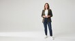 © Shimul Azad - A full-length portrait of a smiling woman standing against a plain white background, wearing a dark green blazer, a white t-shirt, blue jeans, and white sneakers, looking at the