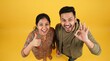 © haris - Happy young couple looking up at the camera, smiling broadly and showing positive hand gestures against a vibrant yellow background.