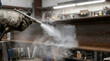 © Sirajum - Industrial worker uses high pressure sandblasting equipment to remove rust and corrosion from steel beam surface in a professional metal fabrication workshop setting with bright lighting