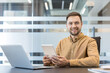 © Liubomir - Digital transformation and remote work, smiling businessman using tablet and laptop in modern office. Professional employee working with technology for business productivity and connectivity.