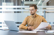© Liubomir - Productivity and remote work, professional man working on laptop and reviewing documents in modern office. Business strategy and data analysis, focused employee managing workflow and checking reports.