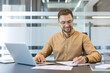 © Liubomir - Professional businessman working on laptop and writing notes in modern office, corporate productivity and career success concept. Focused employee managing tasks and planning strategy at desk.