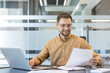 © Liubomir - Professional productivity and corporate workflow, smiling businessman working on laptop and reviewing documents in modern office. Business strategy and data analysis.