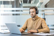 © Liubomir - Remote work and digital communication, man wearing headphones working on laptop in modern office for business productivity and virtual meeting. Professional employee focusing on tasks with technology.