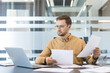 © Liubomir - Workload and deadline pressure, focused businessman reviewing financial documents and analyzing data on a laptop in a modern Office.