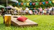 © Ronald Red - Glass of cold beer on cornhole board at outdoor festival. People relaxing in background with bunting flags in park. Summer recreation, leisure activity and fun social gathering event.