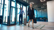 © master1305 - Smiling businesswoman carries a travel bag while colleague pulls a suitcase through airport lobby with upbeat confidence. Corporate arrivals, business mobility and airport transfer concept.
