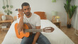 © Krakenimages.com - Man seated on bed playing acoustic guitar and holding smartphone to ear while looking thoughtful in a bedroom building; quiet contemplation.