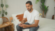 © Krakenimages.com - Man holding smartphone, hand on chin, seated on bed in a tidy bedroom with bedside table, lamp and plants, wearing white tshirt and jeans; quiet contemplation.