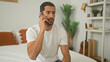 © Krakenimages.com - Man holding smartphone to ear seated on bed in building, wearing white tshirt and jeans with attentive expression; concern solitude.