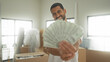 © Krakenimages.com - Man smiling, holding a fanned stack of dollars with right hand while standing among moving cardboard boxes in a building; celebration success joy.
