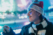 © qunica.com - A young man wearing a Santa hat and scarf smiles while holding a lit sparkler at night. He enjoys a festive outdoor moment with blurred holiday lights and friends.