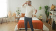 © Krakenimages.com - Man with headphones dancing and swaying with arms raised beside a bed in a building bedroom, wearing sweater and jeans, orange throw visible; joy relaxation.